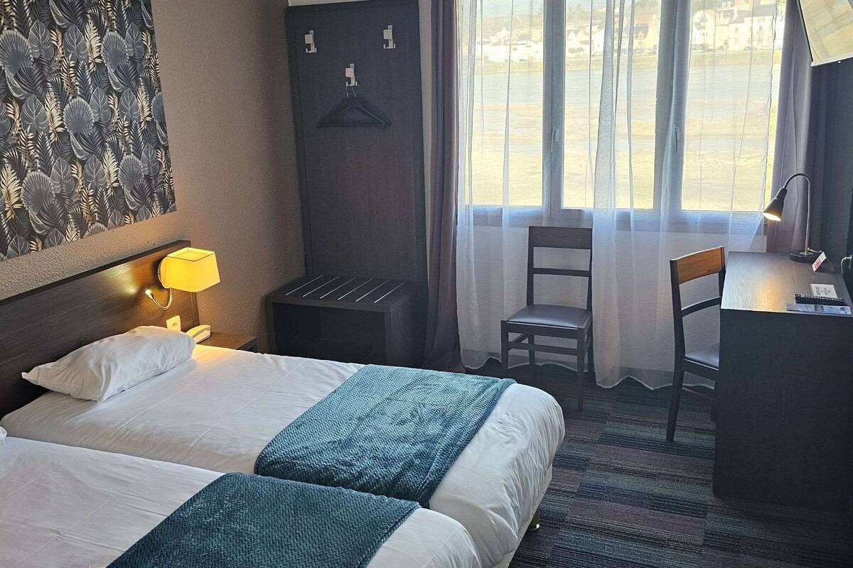 Une chambre d’hôtel avec deux lits, un bureau, une chaise, une lampe et une fenêtre avec des rideaux.