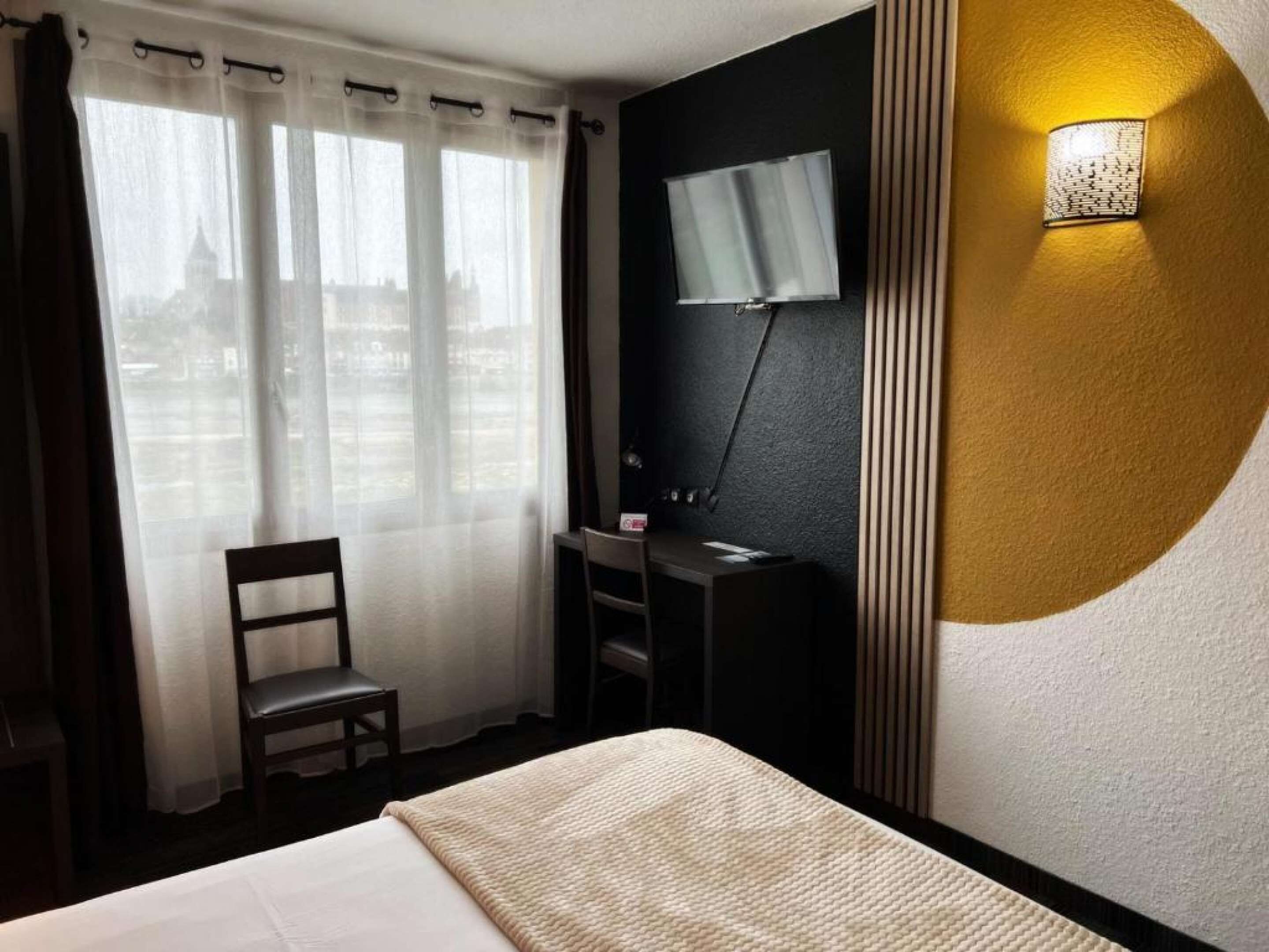 A hotel room with a bed, a desk, a chair, a TV mounted on the wall, and a window with curtains.