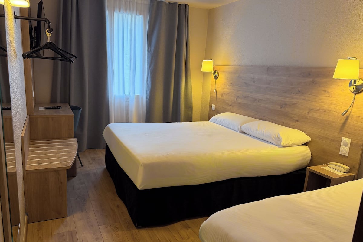 Une chambre d’hôtel comprenant un lit, des lampes de chevet, une table de chevet en bois, un bureau avec une chaise et une fenêtre avec des rideaux.