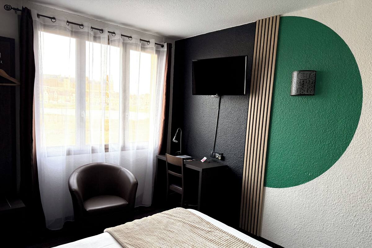 Une chambre d’hôtel avec un lit, une chaise, un bureau avec une lampe, une télévision et un grand mur vert.