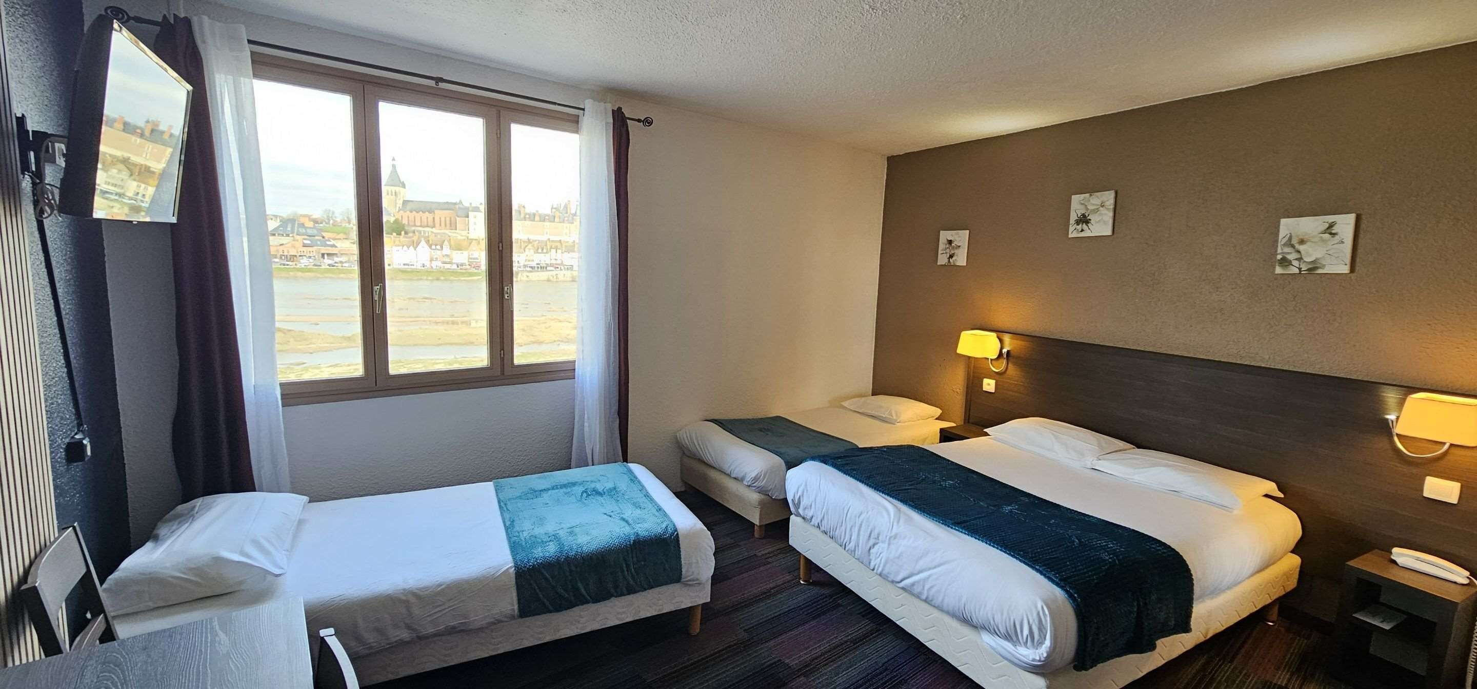 Habitación de hotel con dos camas individuales, un ventanal grande con cortinas, un televisor de pantalla plana en la pared y una mesita con una lámpara.