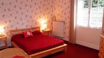 Ein Schlafzimmer mit einem hölzernen Bett, roter Bettwäsche, einem Heizkörper, einem Fenster mit Vorhängen und einem kleinen Nachttisch mit Lampe.