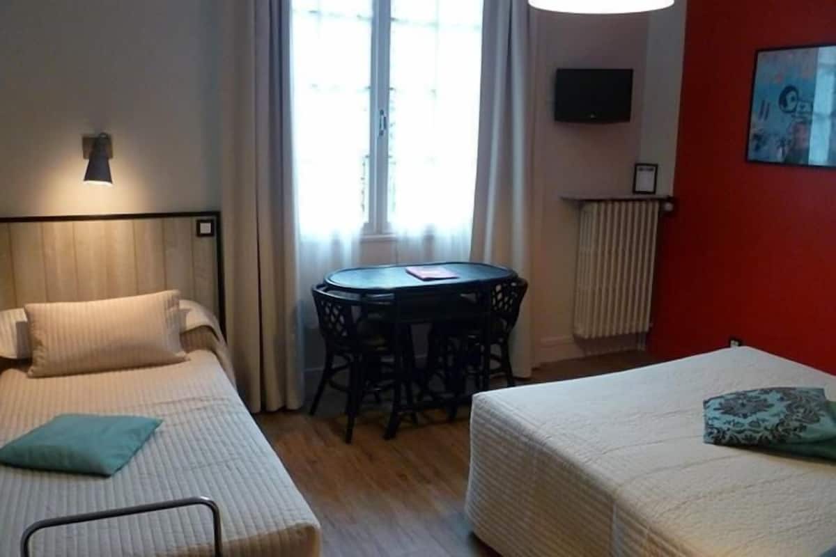 A hotel room with two single beds, a small round table, a TV on the wall, and a window with curtains.