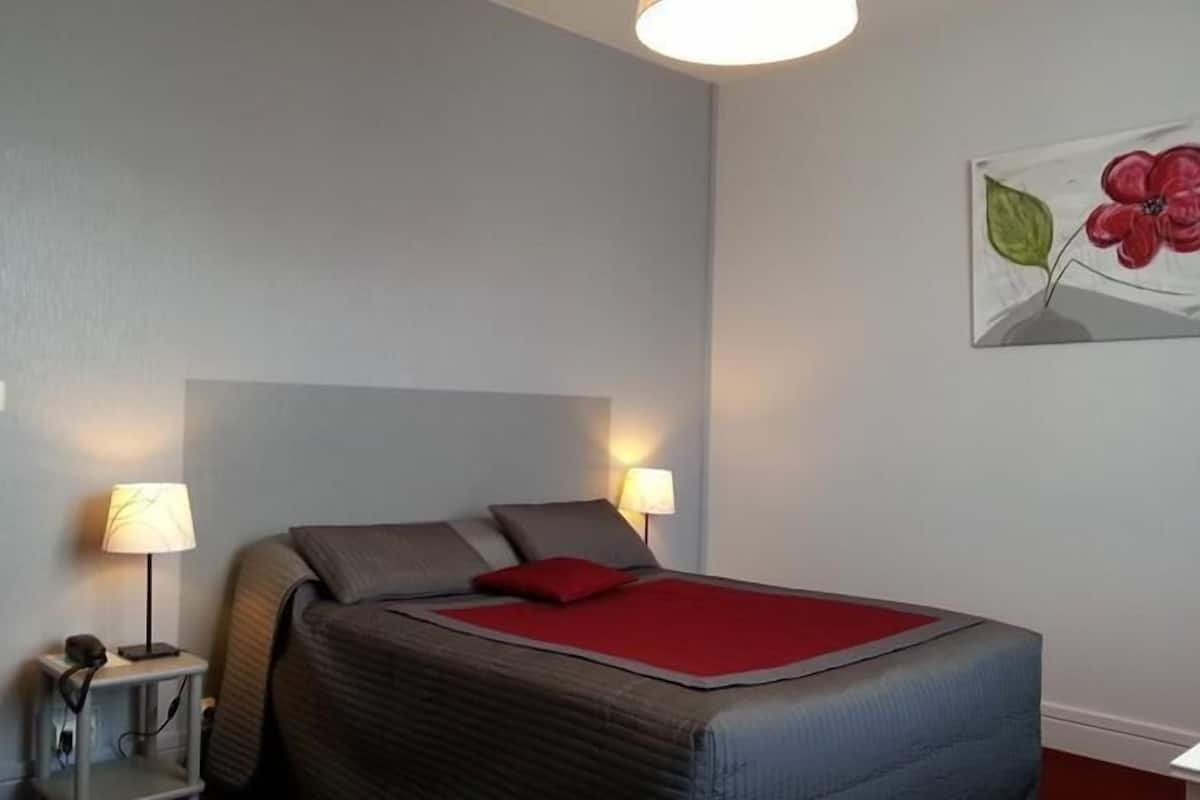 A bedroom with a bed, two bedside lamps, a red throw blanket, and a wall painting.