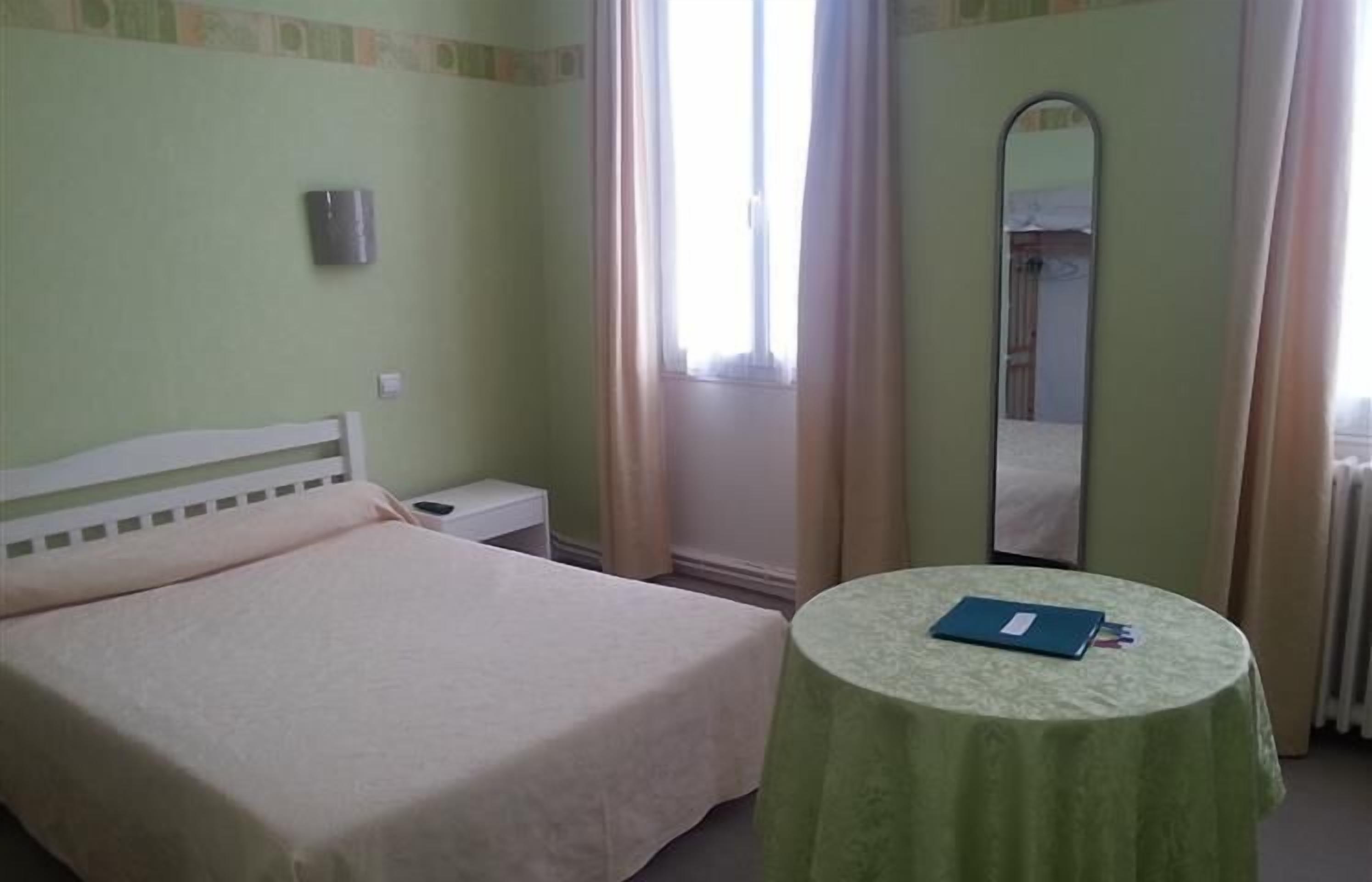 Um quarto com uma cama, mesa de cabeceira, uma mesa redonda com um livro, espelho e janela com cortinas.