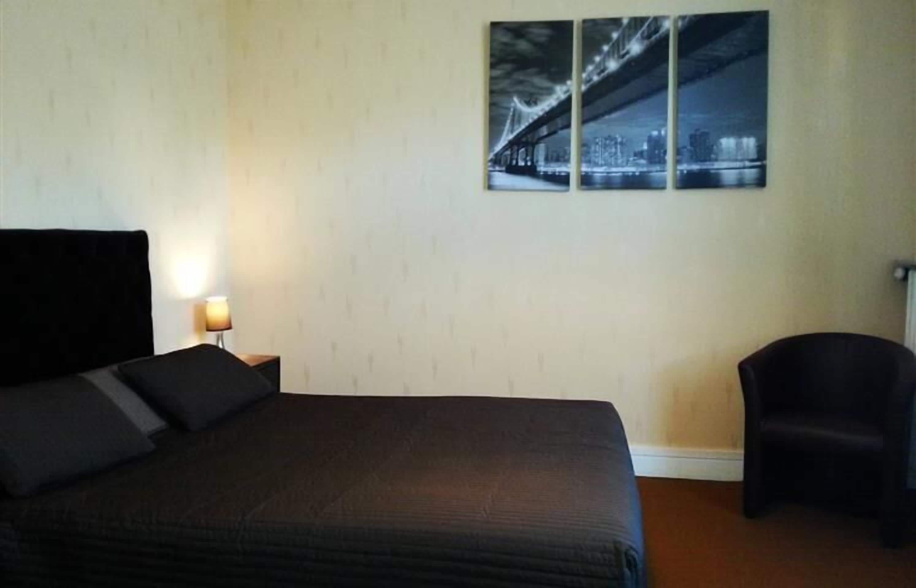 A hotel room with a large bed, a nightstand with a lamp, a chair, and a wall-mounted picture of a bridge.