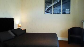 A hotel room with a large bed, a nightstand with a lamp, a chair, and a wall-mounted picture of a bridge.