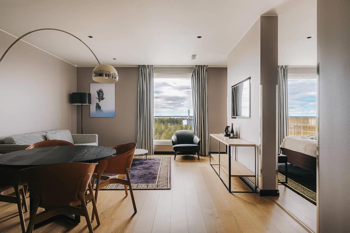 Ein modernes Hotelzimmer mit Essbereich, einer Couch, einem Sessel, einem Schreibtisch, einem Bett und einem großen Fenster mit Aussicht.