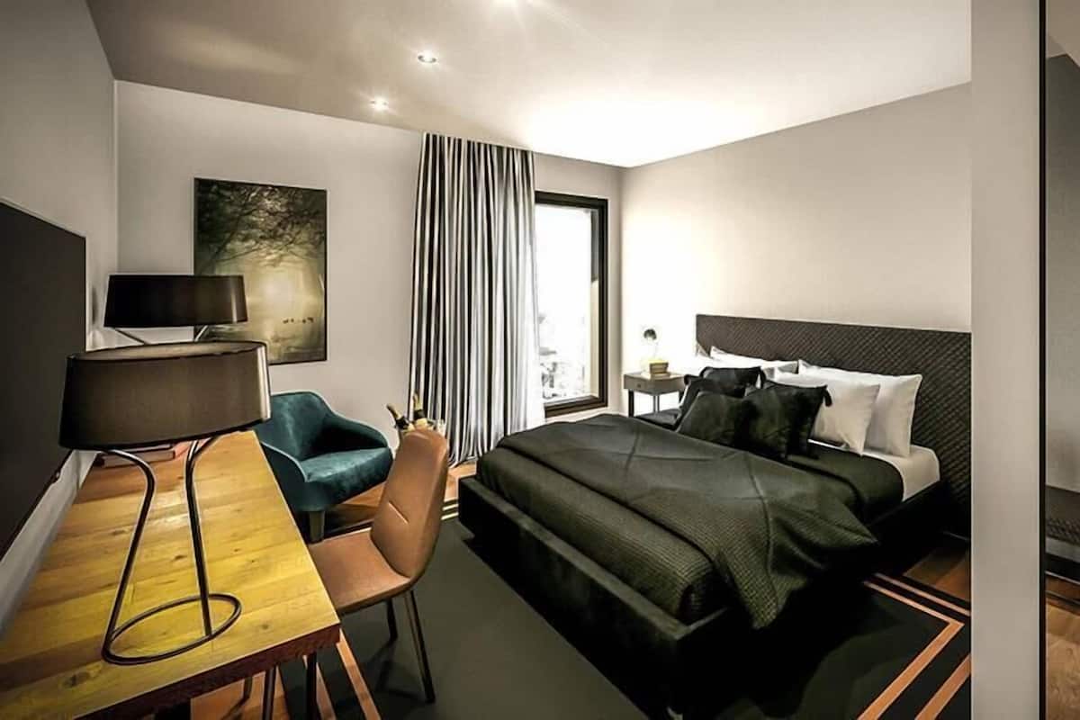 Ein modernes Hotelzimmer mit Bett, Schreibtisch mit Lampe, Stuhl, großem Fenster mit Vorhängen und einem Gemälde an der Wand.