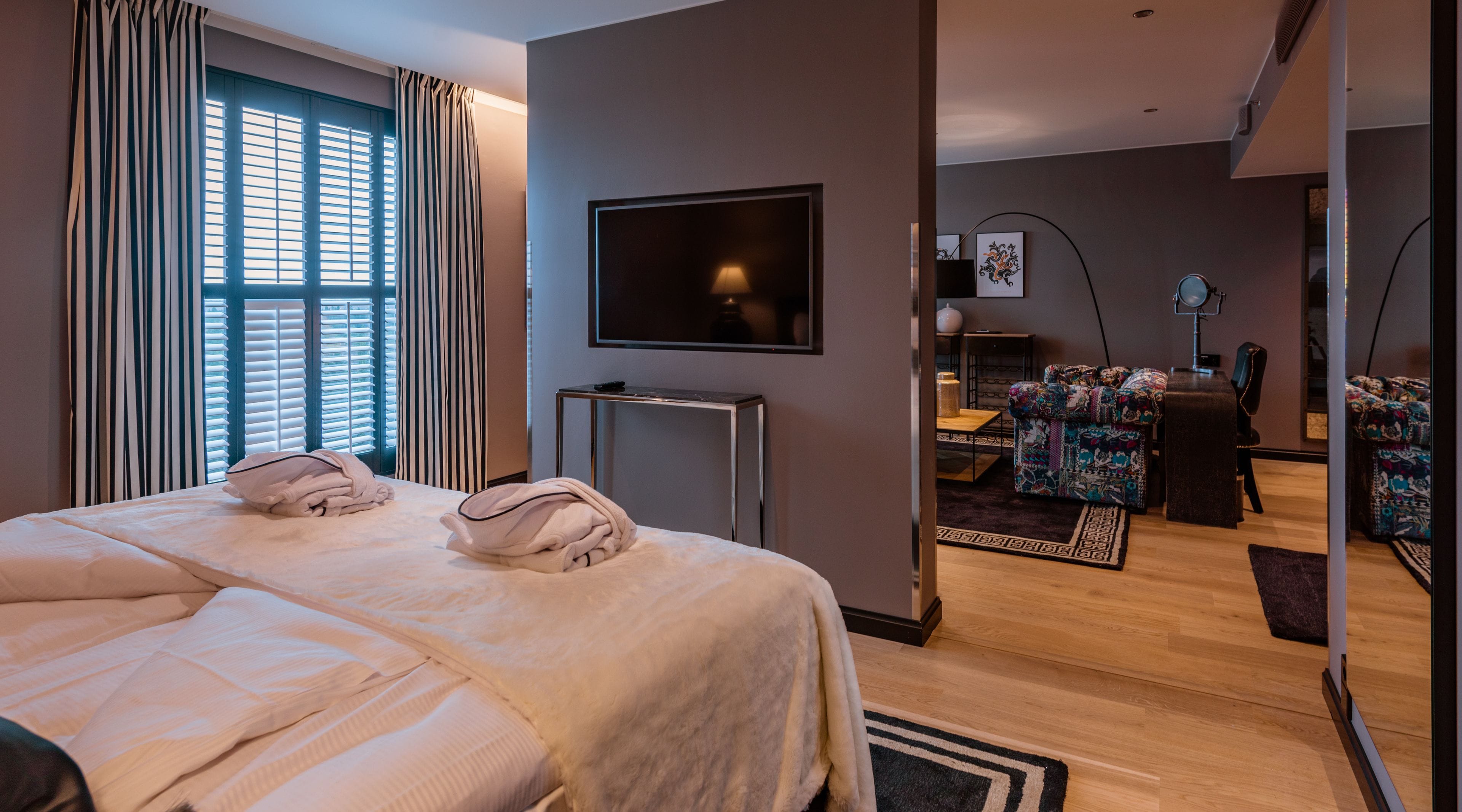 A hotel room with a large bed, a flat-screen TV, a desk with a chair, and a patterned armchair.