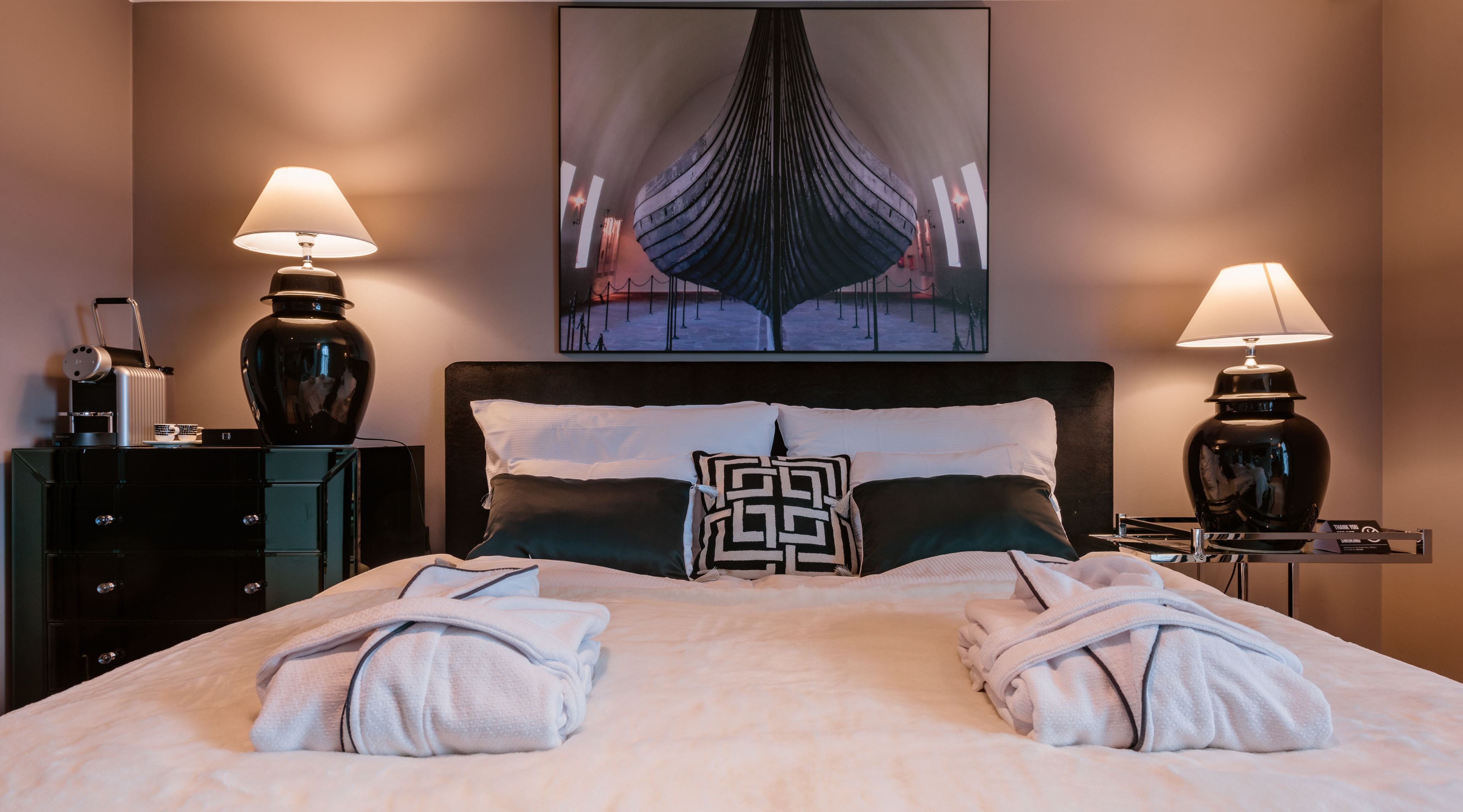 Een netjes opgemaakt bed met handdoeken, twee bedlampen, een hoofdbord met een decoratief kussen, een schilderij boven het bed en een dressoir met een lamp en een klein tafeltje.