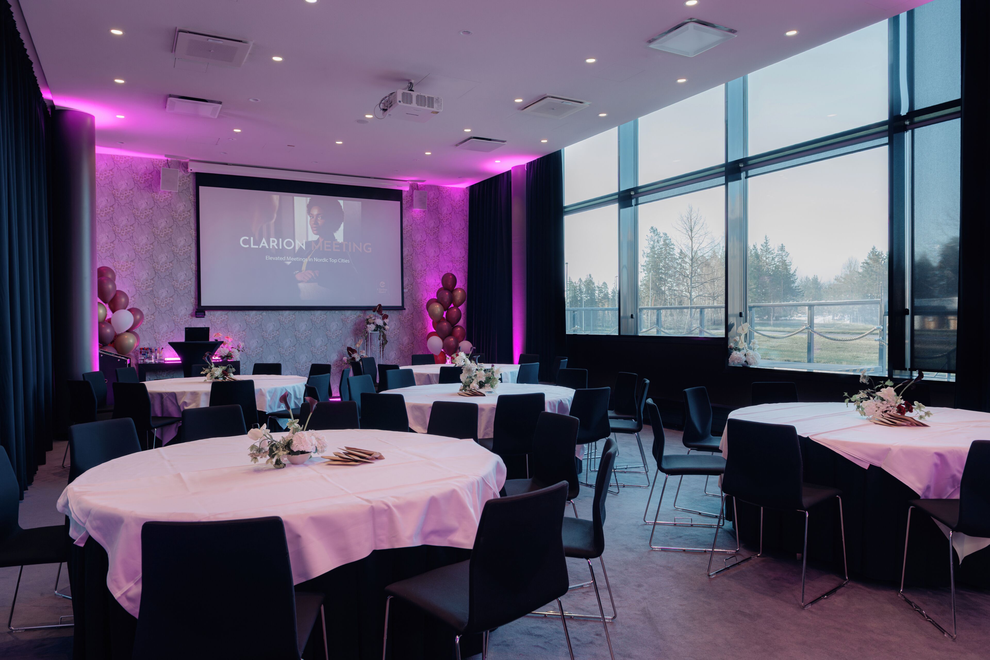 A conference room with round tables, chairs, a projector screen, and a large window.