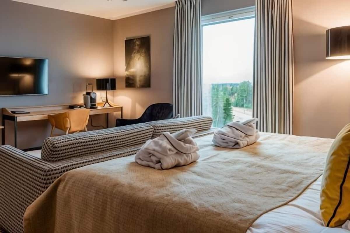 Ein Hotelzimmer mit einem Bett, zwei Nachttischen, einem Schreibtisch mit Stuhl, einem Fernseher und einem Fenster mit Vorhängen.