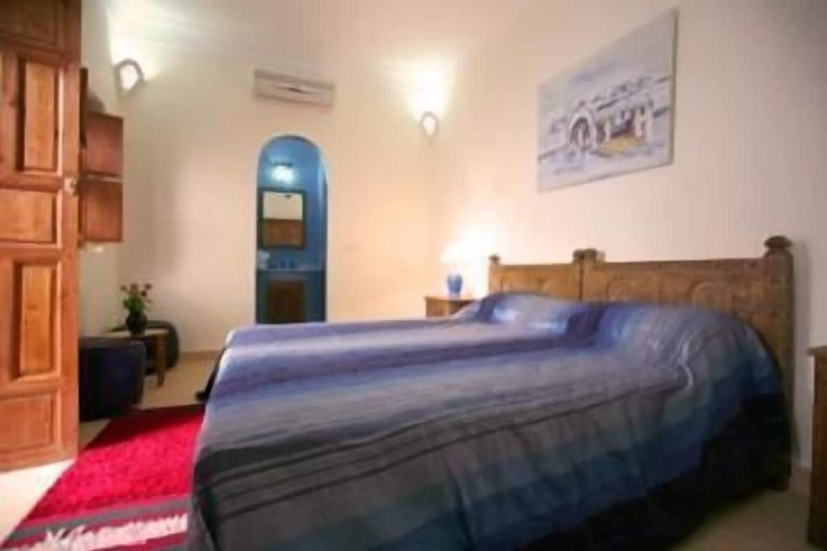 Quarto com cama, mesa de cabeceira, abajur, guarda-roupa de madeira e um quadro na parede.