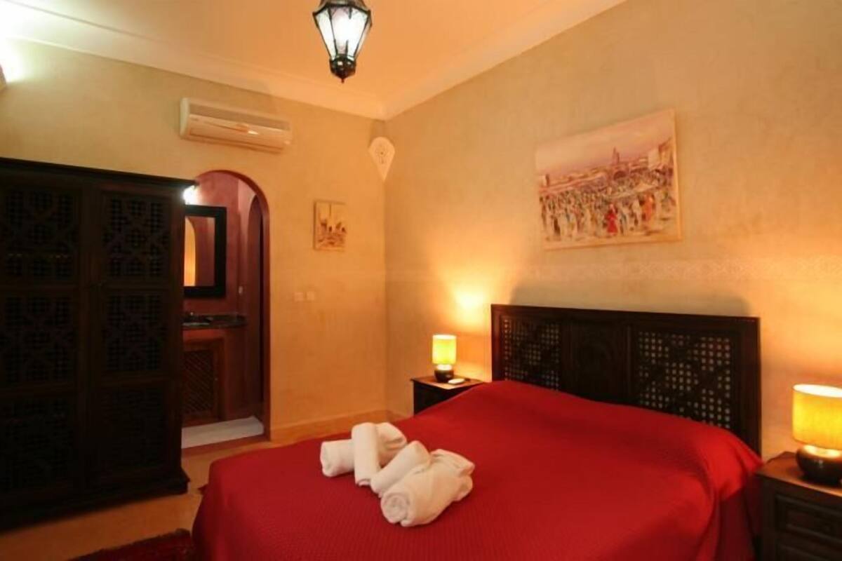 Quarto de hotel com cama vermelha, duas luminárias de cabeceira, um quadro na parede e um aparelho de ar condicionado.