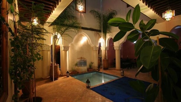 Un área de piscina con suelo de baldosas, columnas arqueadas y plantas en macetas.