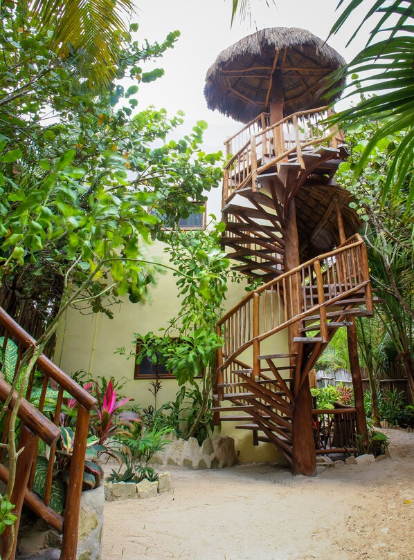 Un escalier en colimaçon en bois mène à une construction au toit de chaume, nichée au cœur d’une végétation luxuriante.