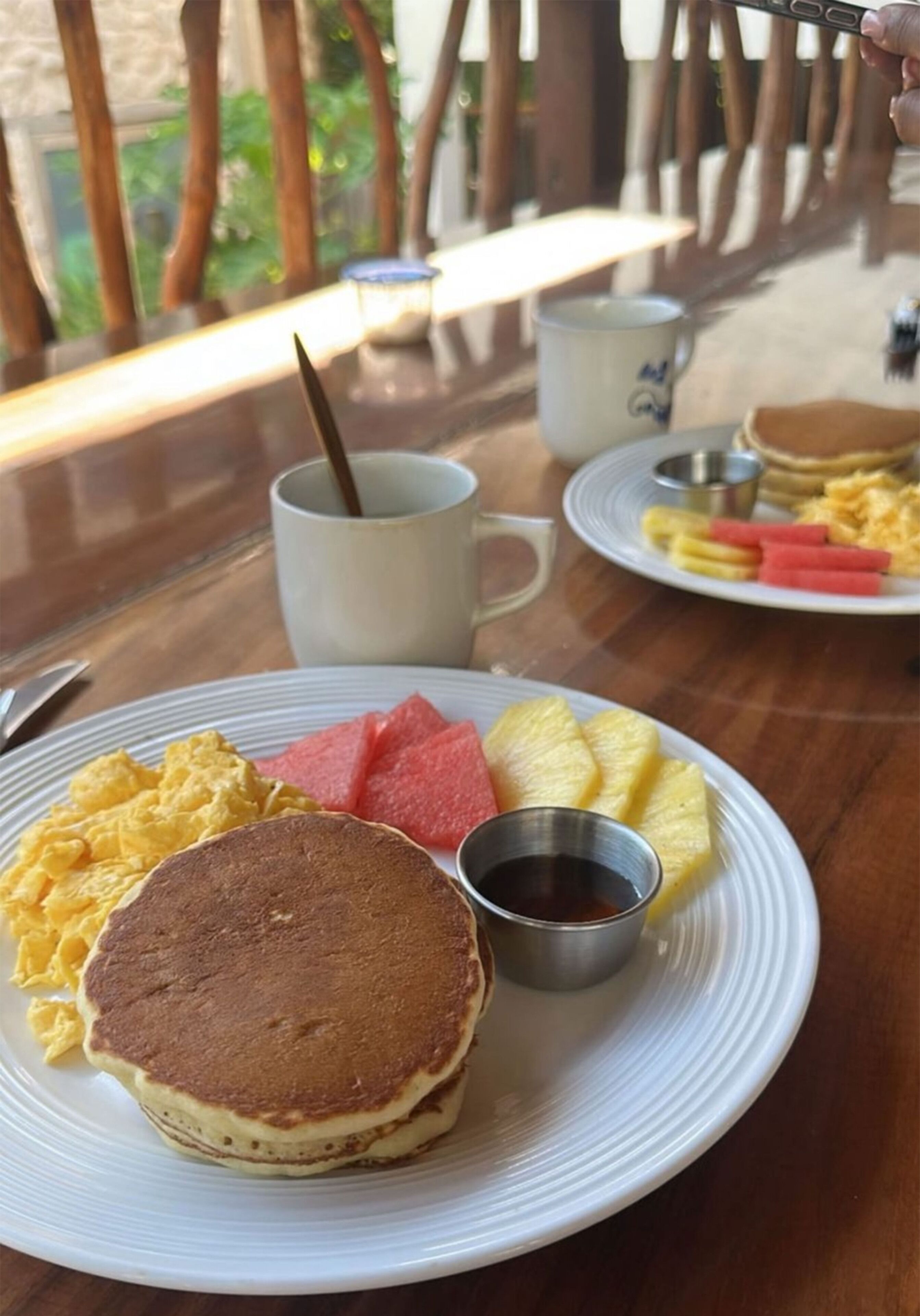Une table dressée pour le petit-déjeuner avec des crêpes, des œufs brouillés, de la pastèque, de l’ananas et une tasse de café.