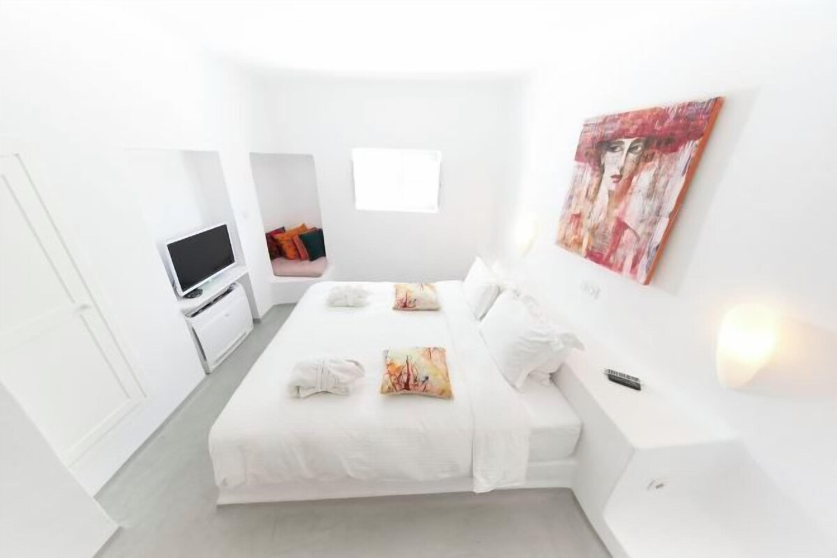A white hotel room with a bed, a TV, a desk, and a painting on the wall.