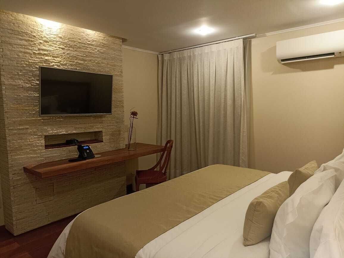 A hotel room with a bed, a TV mounted on a textured wall, a desk with a chair, and an air conditioning unit.