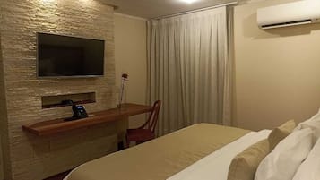 A hotel room with a bed, a TV mounted on a textured wall, a desk with a chair, and an air conditioning unit.