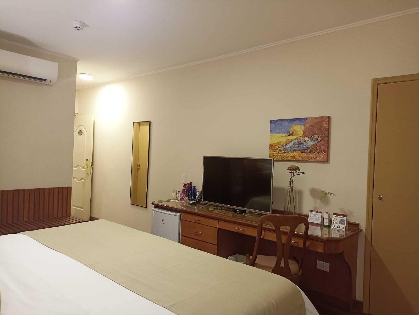 A hotel room with a bed, a desk with a computer, a chair, a television, a mirror, and a painting on the wall.