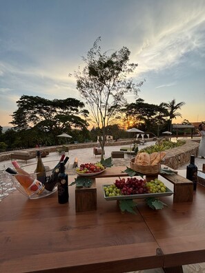 An outdoor dining area with a table set for a meal, featuring a variety of fruits and bottles of wine.