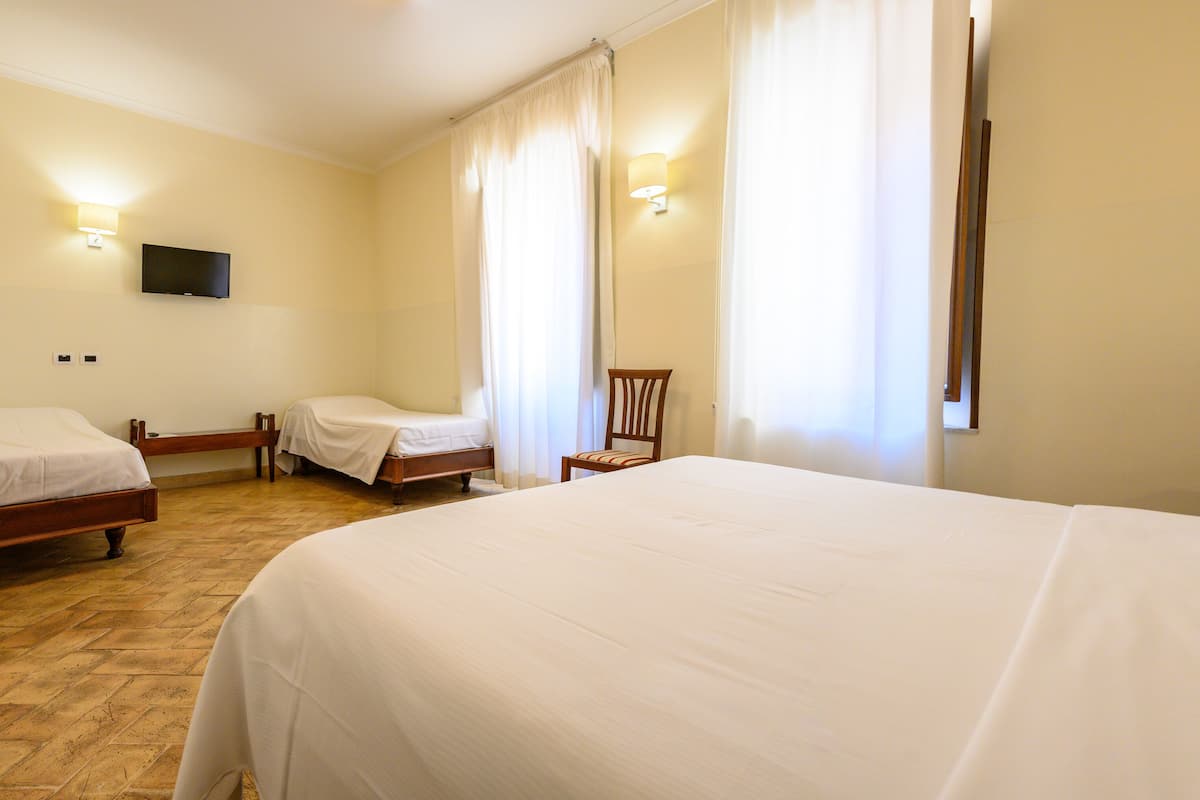 A hotel room with two single beds, a wooden chair, a flat-screen TV mounted on the wall, and a large window with curtains.