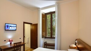 A hotel room with a bed, a desk with a chair, a TV mounted on the wall, and a window with curtains. - Casa I Cappuccini (Rome)
