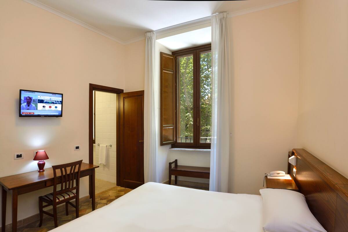 A hotel room with a bed, a desk with a chair, a TV mounted on the wall, and a window with curtains.