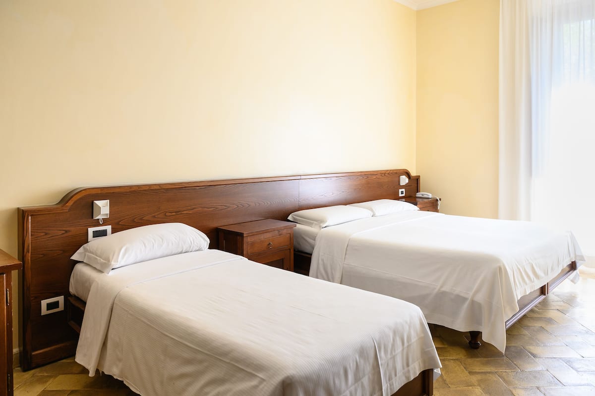 A hotel room with two single beds, wooden headboards, bedside tables, and a large window with curtains.