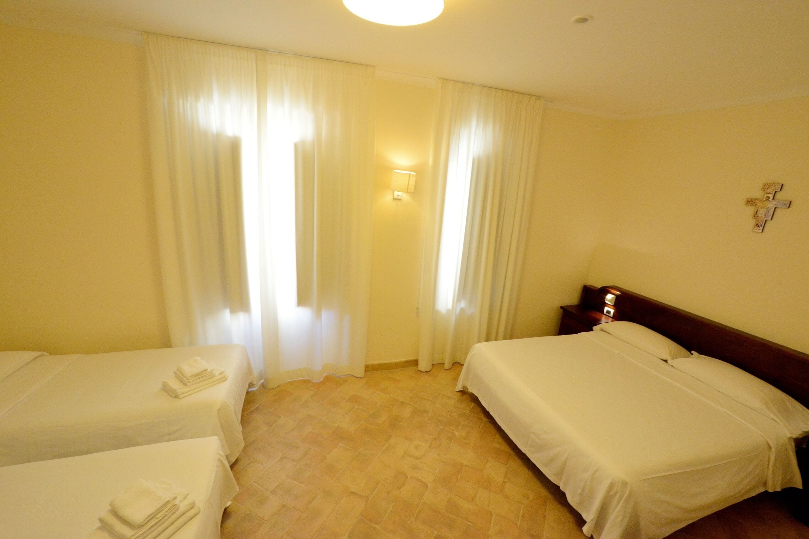 A hotel room with two single beds, a nightstand, a wall-mounted light, and a window with curtains.