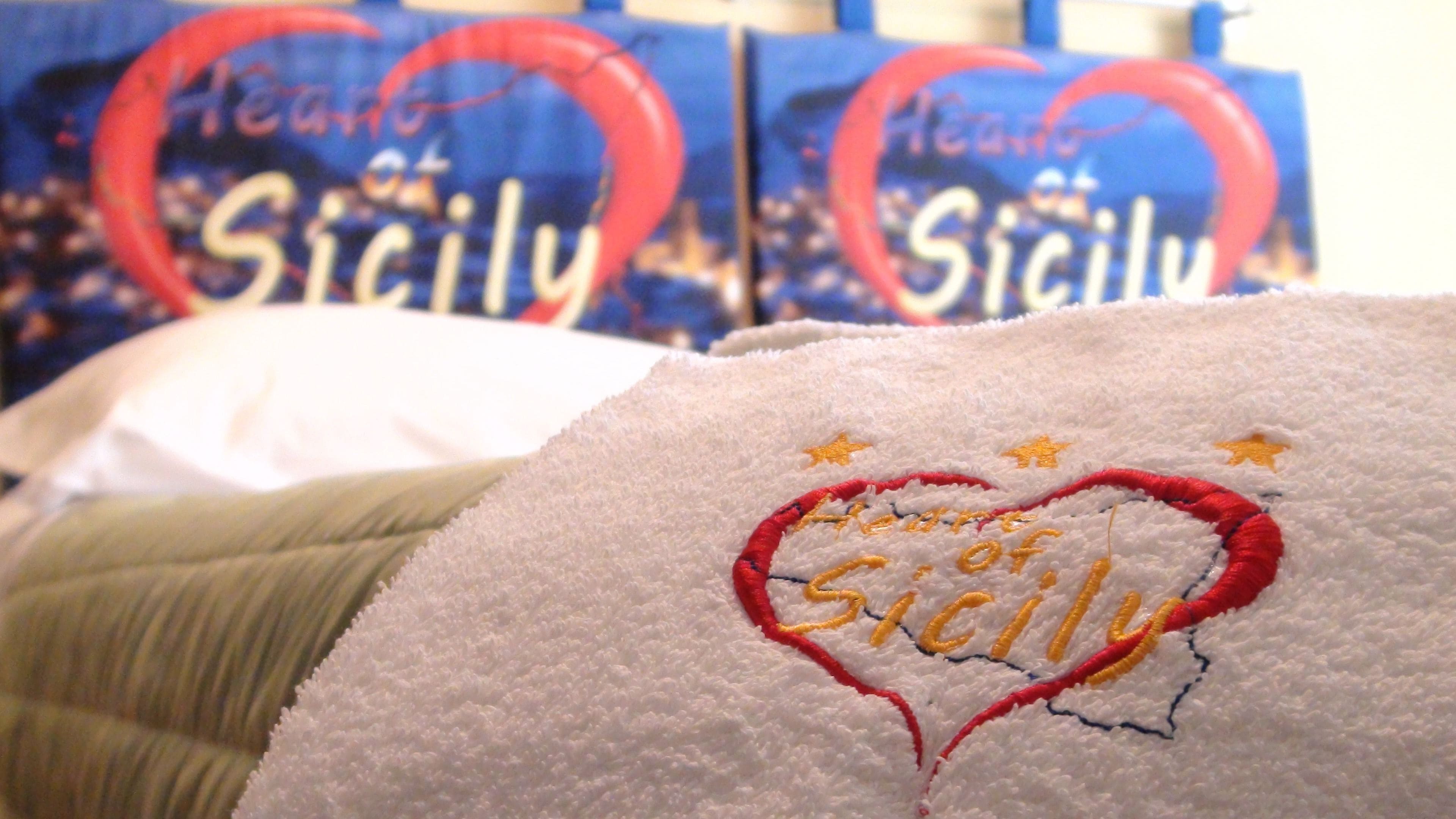 En närbild på en handduk med broderad text och ett hjärtmotiv, och i bakgrunden syns lådor med texten ”New York of Sicily”.
