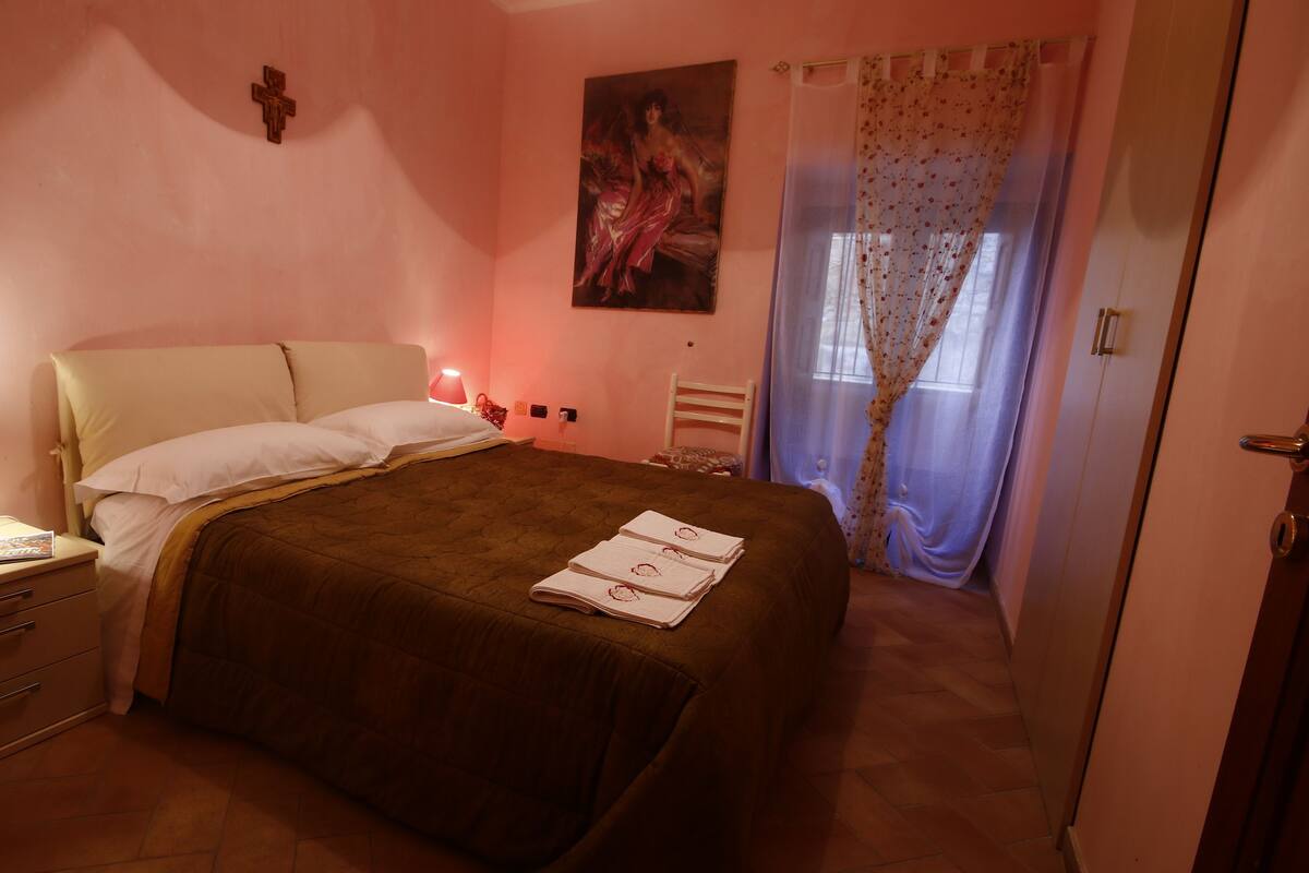 A bedroom with a bed, bedside table with a lamp, a cross on the wall, and a painting.