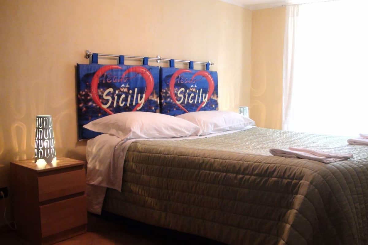 A neatly made bed with a green quilt, a bedside lamp, and two heart-shaped pillows with "Heart of Sicily" text.