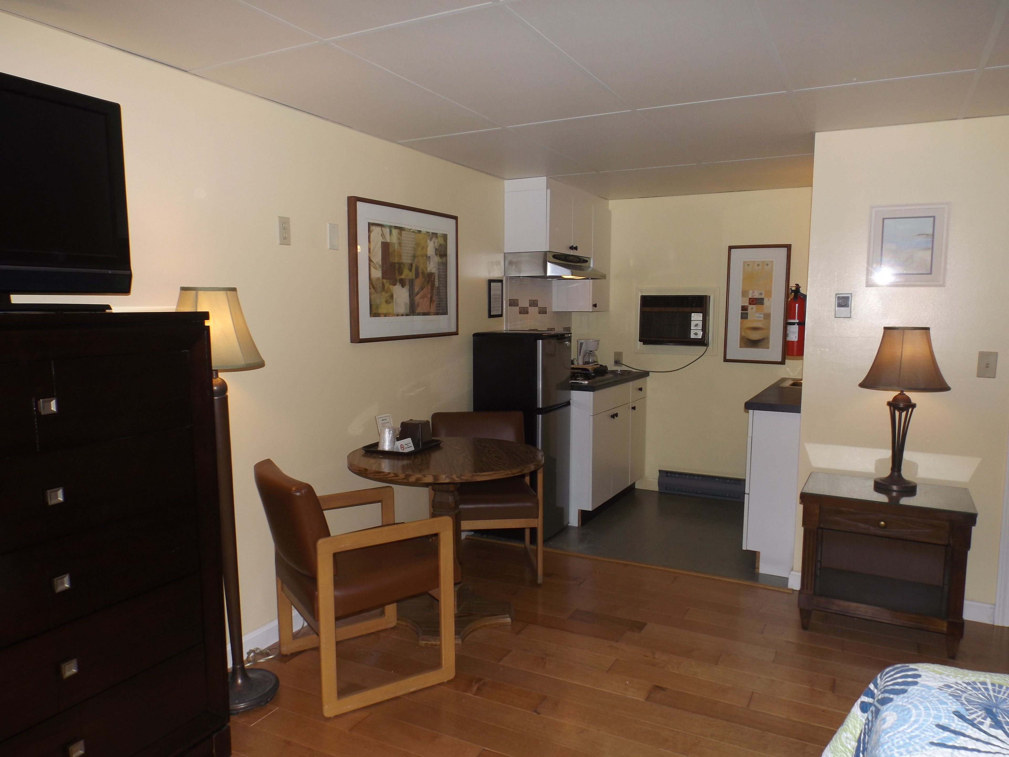 Une chambre d’hôtel compacte comprenant un coin cuisine, une télévision, une chaise en bois, une petite table et un lit recouvert d’une courtepointe à motifs.