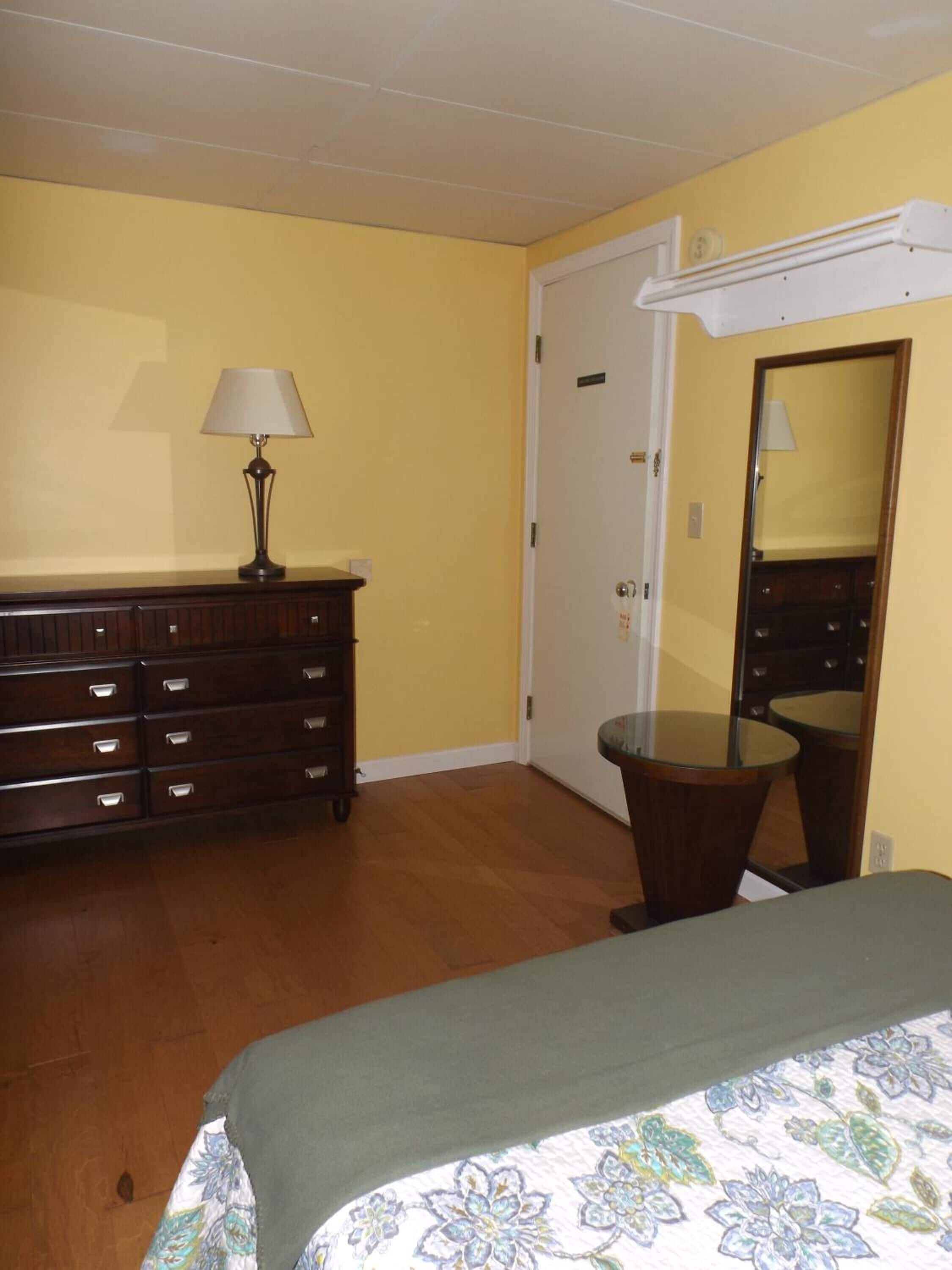 A bedroom with a wooden dresser, a lamp, a bed with a floral bedspread, a round table, and a mirror.