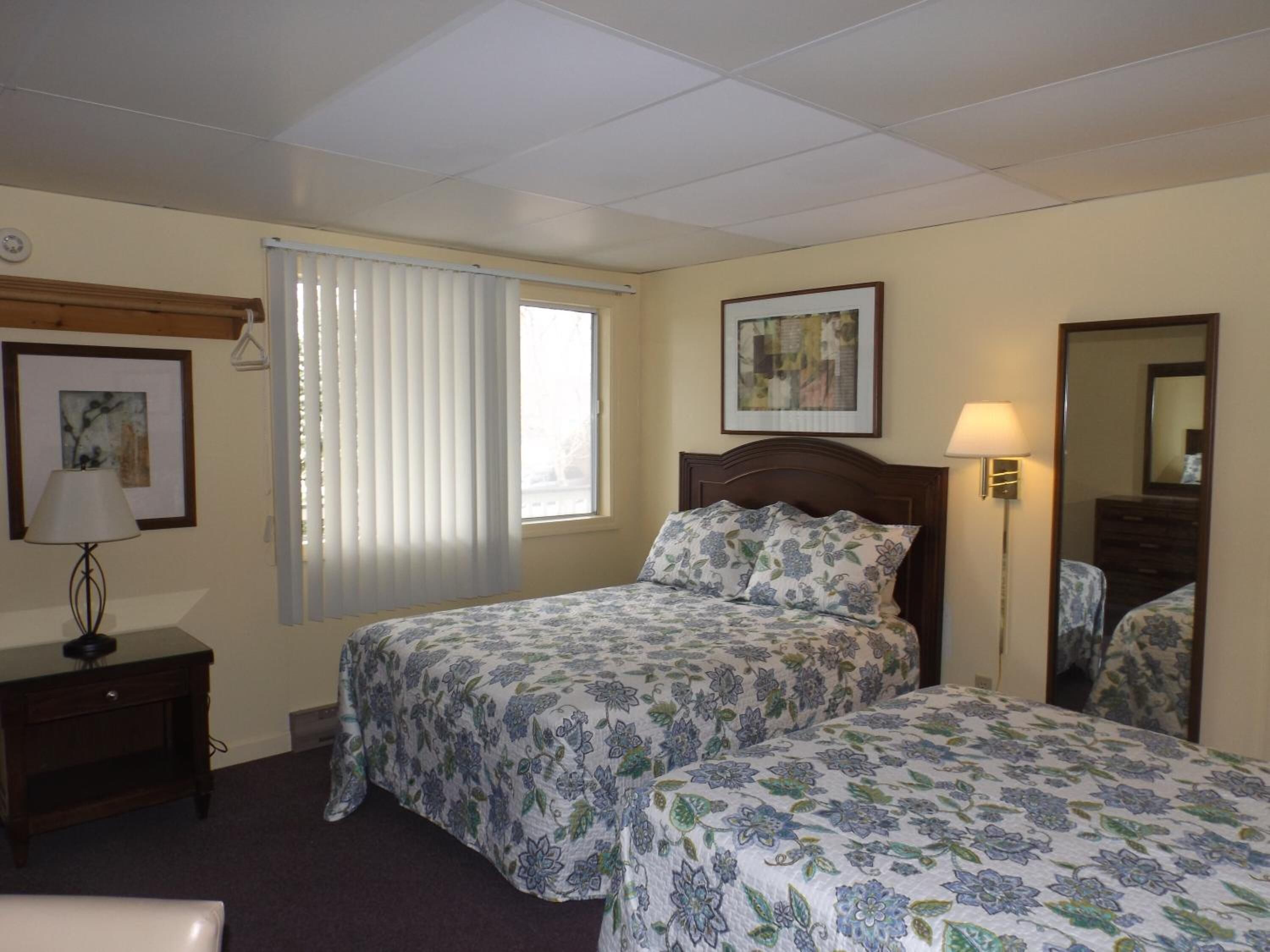 Habitación de hotel con dos camas, una mesita de noche con lámpara, una ventana con persianas y un cuadro en la pared.