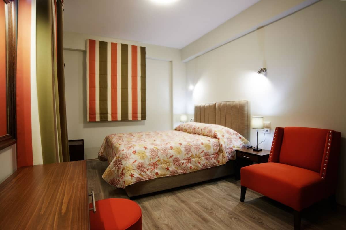 A hotel room with a bed, a red chair, a wooden table, and a striped wall hanging.
