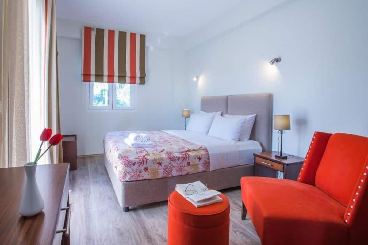 A hotel room with a bed, two bedside tables, a red armchair, a wooden table with a vase, and a window with curtains.