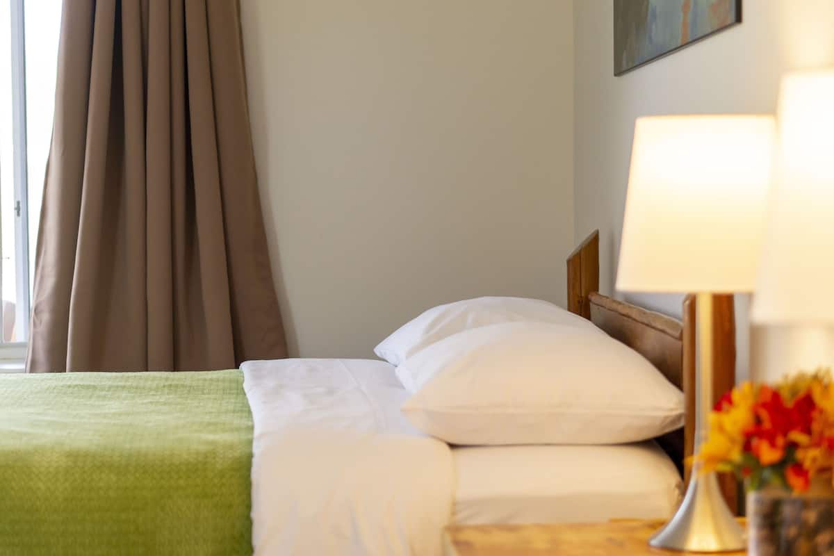 A neatly made bed with white linens and a green blanket, a bedside lamp, and a vase with flowers.