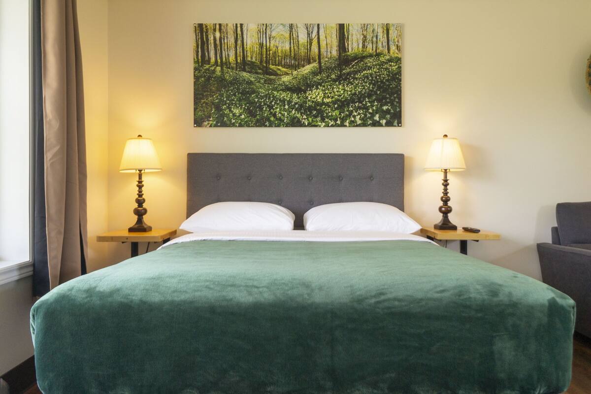 A neatly made bed with a green bedspread, two bedside lamps, and a forest-themed painting above the bed.