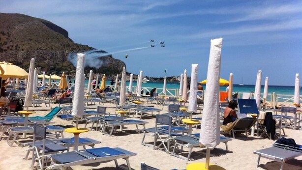 A beach with rows of sun loungers and umbrellas, a clear sky, and a distant aircraft formation. - B&B Teatro (Palermo)
