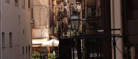 A narrow cobblestone street flanked by buildings with balconies, a clock tower, and a street lamp.