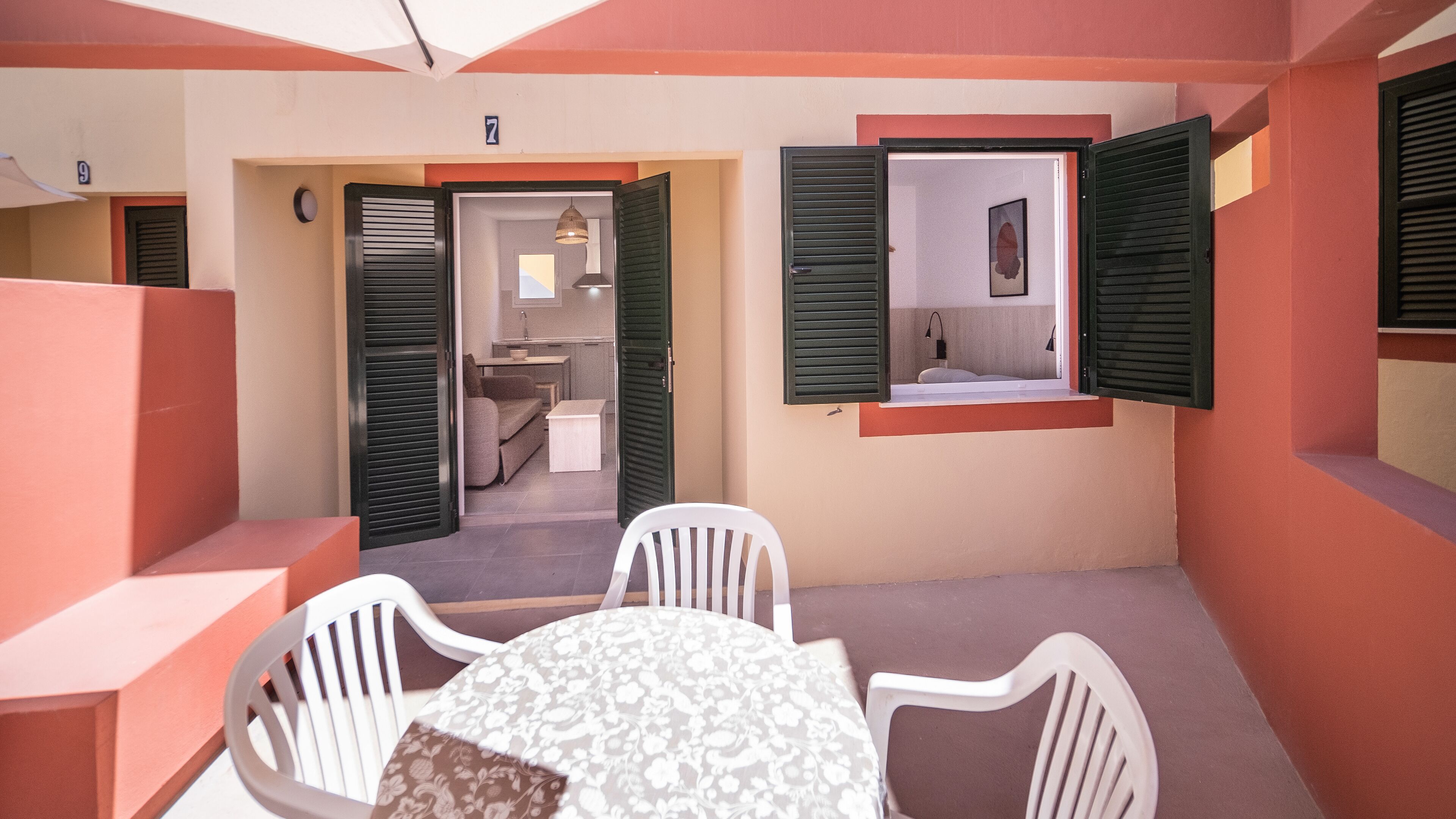 Un pequeño balcón cerrado con una mesa y sillas, una ventana con contraventanas y vista al cuarto contiguo.