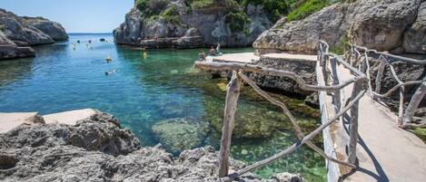 Zona costera rocosa con agua cristalina, un paseo marítimo de madera y vista al mar.