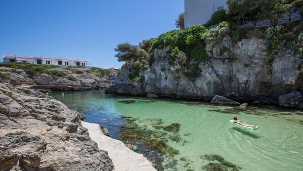 Escena costera con agua cristalina, costa rocosa y un edificio con balcones.