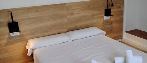 A neatly made bed with white linens and towels, a wooden headboard, and two wall-mounted lamps.