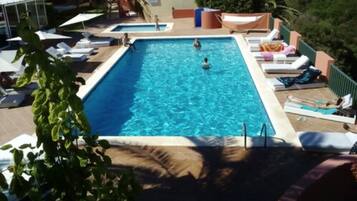 Una piscina rectangular con una persona nadando, rodeada de sillas de descanso y sombrillas.