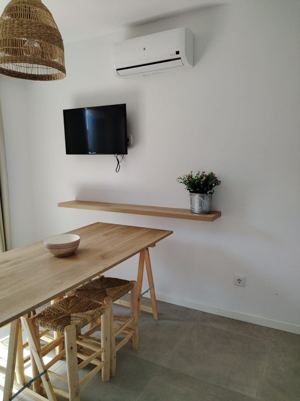 Una habitación minimalista con una mesa de madera, una silla de mimbre, un televisor montado y una pequeña planta en maceta.